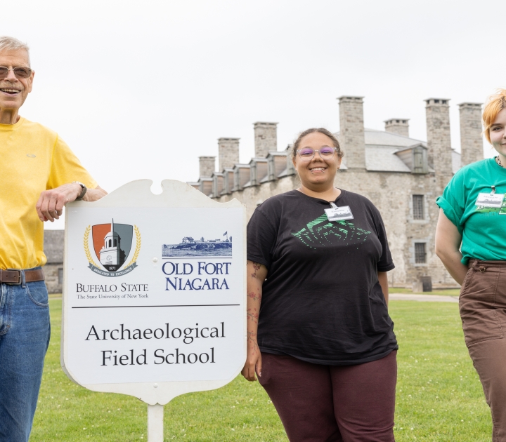 Three representatives standing by a sign reading Buffalo State Archaeological Field School sign at Old Fort Niagara